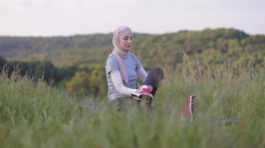 Happy sportswoman in hijab and activewear stretching leg muscles while sitting on mat in the park