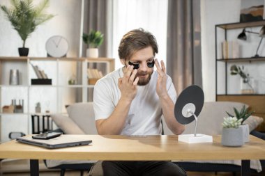 Young bearded Caucasian man applying black eye patches at home