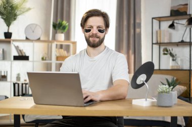 Caucasian man, with black collagen eye patches, sitting at the table at home and working on laptop