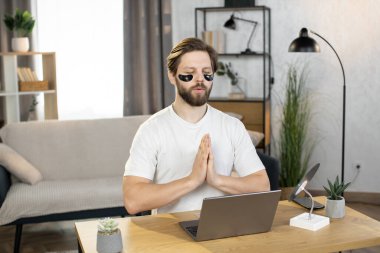 Caucasian man meditating at home after work on laptop, sitting at the table with namaste gesture