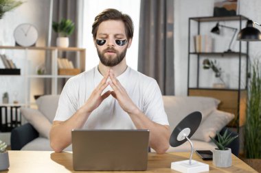 Caucasian man meditating at home after work on laptop, sitting at the table with namaste gesture