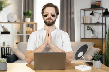 Caucasian man meditating at home after work on laptop, sitting at the table with namaste gesture