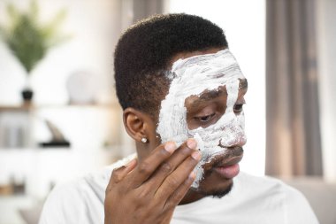 african american guy applying white facial mask on face and looking on camera