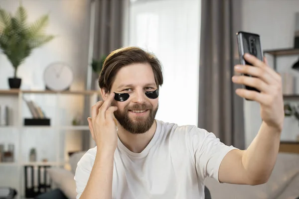 Caucasian man, with black hydrogel patches under eyes, sitting at home and talking on the phone — Stock Photo, Image