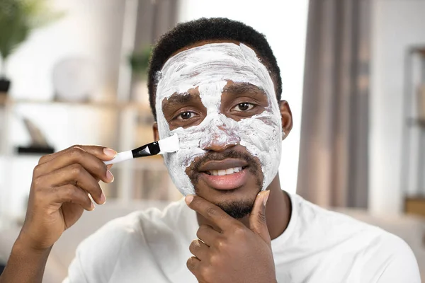 Handsome african american applying white clay mask on his face with brush — Stock Photo, Image