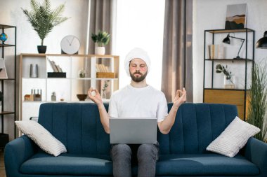 Kafasında havlu ve yamalar olan bir adam, kanepeye oturmuş, gözleri kapalı meditasyon yapıyor.