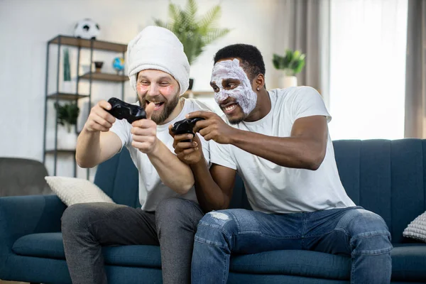 Мультиэтнические мужчины делают процедуры по уходу за кожей, играют в видеоигры с помощью джойстиков — стоковое фото