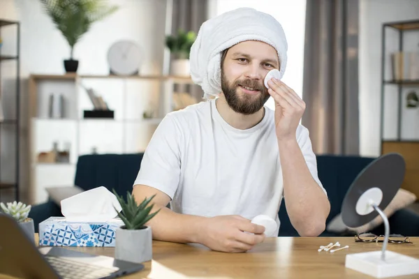 Caucasian man in towel on his head wiping his face with round cotton pad in his hand. — Stock Photo, Image
