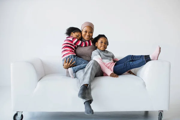 Happy african american muslim mother resting with daughters on sofa