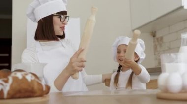 Anne ve küçük kız aşçı şapkası takıp gülüyor ve merdanelerin üzerinde kavga ediyorlar.