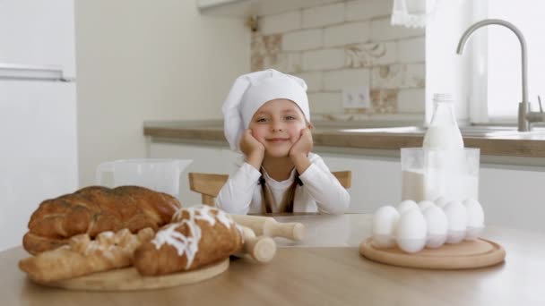 Pequeña chica blanca alegre en ropa casual ligera sentada en una mesa de cocina de madera — Vídeos de Stock