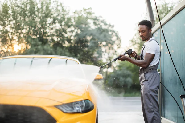 Sarı arabasını yıkayan koruyucu tulumlu yakışıklı Afrikalı genç. — Stok fotoğraf