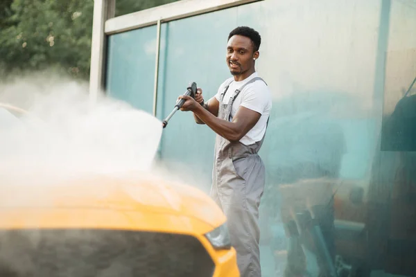 Jóképű afrikai fiatalember védőruhában, mossa a sárga kocsiját. — Stock Fotó