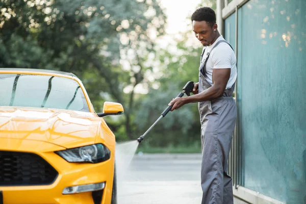 Africano joven enfocado en el hombre en la ropa de trabajo lavar el coche amarillo con una pistola de agua —  Fotos de Stock