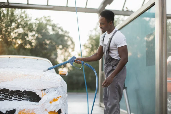 Afrykanin myje swój żółty samochód pianką w samoobsługowej myjni samochodowej na zewnątrz. — Zdjęcie stockowe