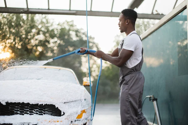 Afrykanin myje swój żółty samochód pianką w samoobsługowej myjni samochodowej na zewnątrz. — Zdjęcie stockowe