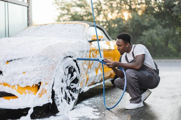 Joven hombre africano con chorro de alta presión rociando la espuma en la rueda del coche en la estación de servicio de lavado de coches —  Fotos de Stock