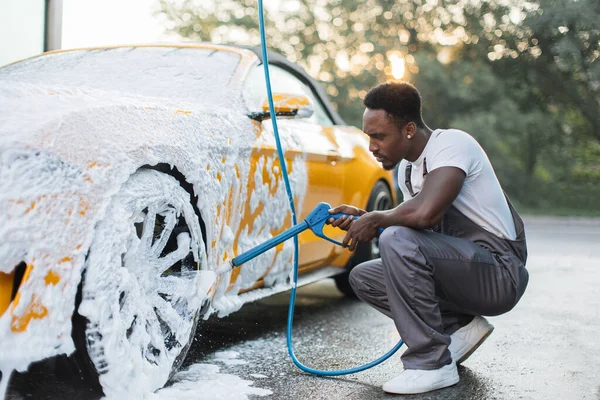 Yüksek basınçlı jeti olan genç bir Afrikalı araba yıkama istasyonunda araba tekerine köpük püskürtüyor. — Stok fotoğraf