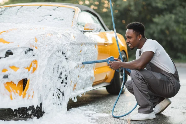 Fiatal afrikai férfi mosás kerekek a luxus autó permetezés a hab nagy nyomással . — Stock Fotó