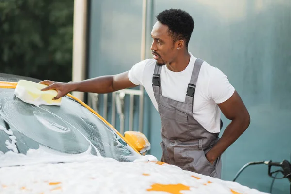 Çekici Afrikalı Amerikalı genç adam arabasını temizleme köpüğü ve sarı süngerle yıkıyor.. — Stok fotoğraf