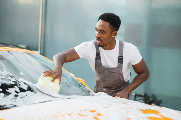 Joven atractivo hombre afroamericano lavando su coche con espuma limpiadora y esponja amarilla. — Foto de Stock