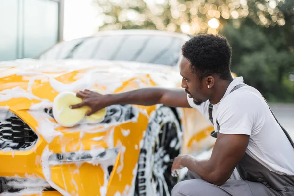 Jóképű afrikai sötét bőrű férfi védő overálban, mossa a sárga autó fényszórók — Stock Fotó