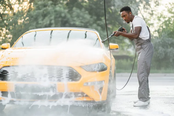 Koruyucu tulumlu Afrikalı bir adam arabasını yüksek basınçlı su hortumuyla elle yıkıyor. — Stok fotoğraf