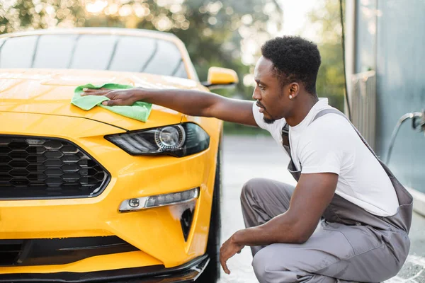 Gri tulumlu, beyaz tişörtlü, araba kaputunu temizleyen gülümseyen Afrikalı genç bir adamın portresi. — Stok fotoğraf