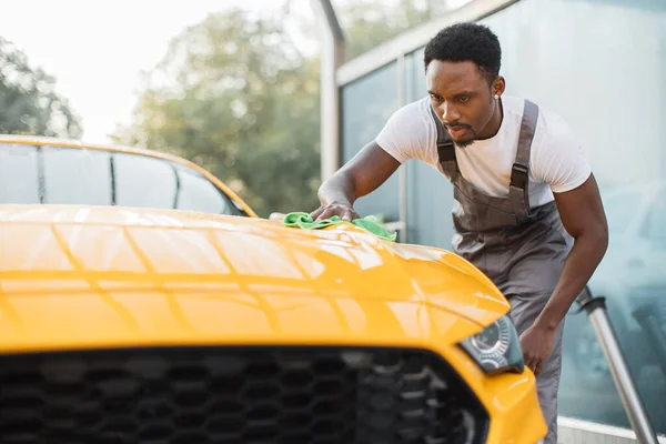 Sarı araba kaputunu temizleyen yakışıklı sakallı genç Afrikalı işçi. — Stok fotoğraf
