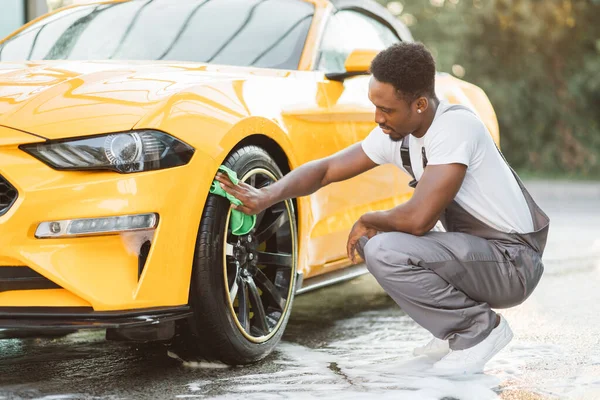 Guapo africano chico limpiando rueda de coche de lujo moderno con paño de microfibra verde —  Fotos de Stock