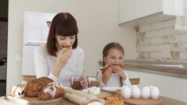 Menina bonito e sua mãe com rostos untados de farinha estão comendo croissants e bebendo cacau — Vídeo de Stock