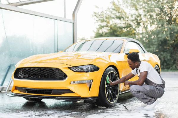 Tampan Afrika pria menyeka dengan kain hijau roda mobil selama proses mencuci luar ruangan. — Stok Foto