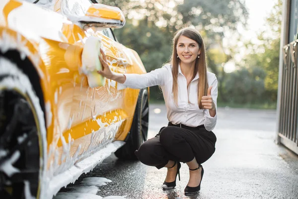 Wanita muda dalam pakaian bisnis, kemeja dan celana menggunakan spons kuning untuk membersihkan pintu mobil — Stok Foto