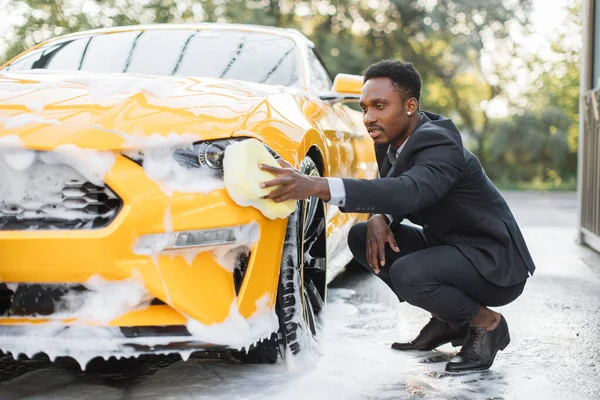 身穿西服的非洲人，用黄色海绵和肥皂洗洗他的黄色汽车前灯. — 图库照片