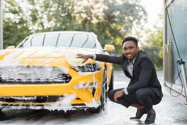Hombre afroamericano guapo en traje de negocios lavando capucha de coche — Foto de Stock