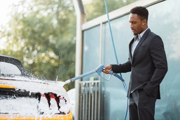 Portret przystojnego brodatego młodego afrykańskiego biznesmena myjącego swój żółty samochód pianką — Zdjęcie stockowe