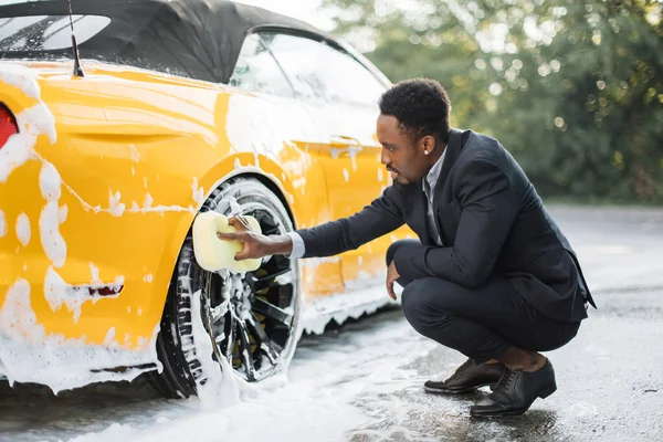 Oldalnézet jóképű fiatal afrikai üzletember használ sárga szivacs tisztítására felnik szabadban. — Stock Fotó