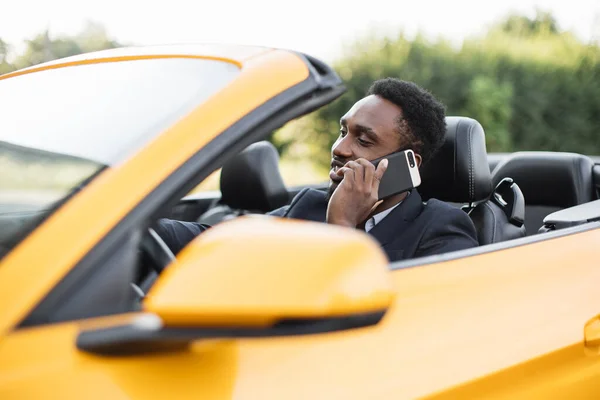 Hombre de negocios africano guapo hablando en el teléfono móvil mientras está sentado en el asiento trasero de un coche deportivo. — Foto de Stock