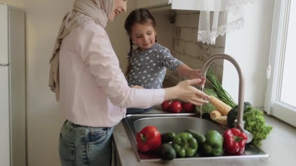 Een jonge mooie moeder in hijab en haar dochtertje wassen groenten onder de kraan met water — Stockvideo