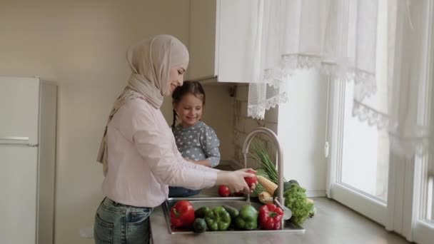 Junge schöne muslimische Mutter und ihre süße Tochter waschen gemeinsam Gemüse in einer Küchenspüle — Stockvideo