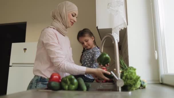 Kleines Mädchen hilft ihrer hübschen muslimischen Mutter beim Gemüsewaschen in der Küche. — Stockvideo