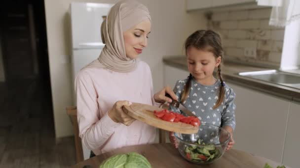 Arabska matka w hidżabie i słodka córeczka rzucająca krojone ogórki do szklanej miski — Wideo stockowe