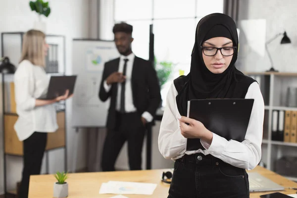 Femme dans le hijab en utilisant le presse-papiers avec des collègues au bureau — Photo