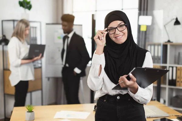 Mujer en hijab y gafas sujetando portapapeles durante la reunión —  Fotos de Stock