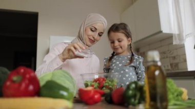 Tesettürlü gülümseyen anne portresi ve küçük beyaz kız salataya tuz ekler.