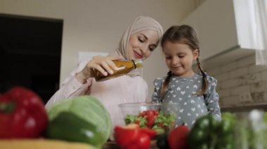 Tesettürlü gülümseyen anne portresi ve küçük beyaz kız salataya tuz ekler.