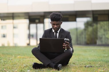 Afro-Amerikalı adam dışarıda dizüstü bilgisayar kullanıyor, ofisin yanındaki çimlerin üzerinde oturuyor.