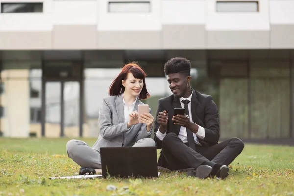 Двоє багаторасових бізнес-колег, сидячи на траві насолоджуючись перервою, використовуючи телефони і показуючи лайки — стокове фото