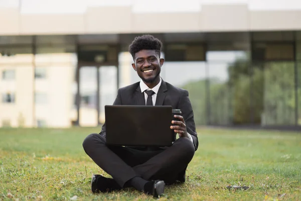 Jeune homme d'affaires africain portant un costume noir assis sur l'herbe avec un ordinateur portable et emporter du café. — Photo