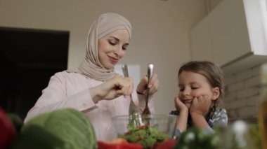 Modern sıcacık evcil mutfakta küçük sevimli kız, genç müslüman anneye sağlıklı yemek pişirmesinde yardım eder.